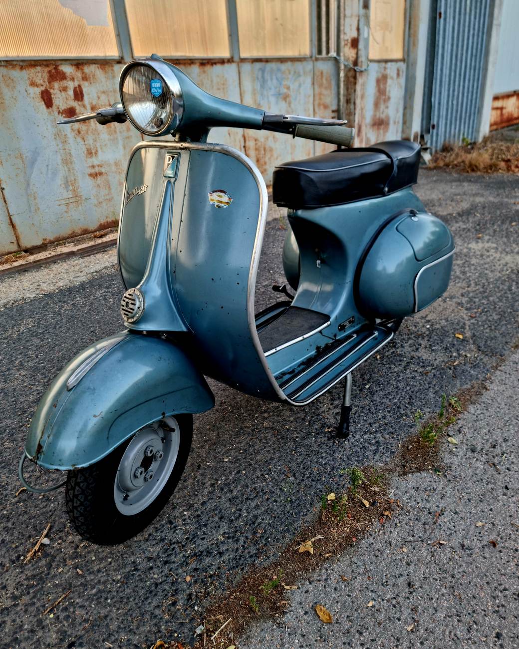 Patinás, kék-szürke klasszikus Vespa háromnegyed nézetben, esti fényben egy kopott ipari fal előtt